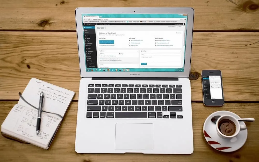 Laptop on a desk with coffee, notebook and an iphone